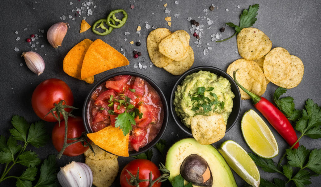 Guacamole, fresh salsa, chips, lime and peppers arranged on a dark table